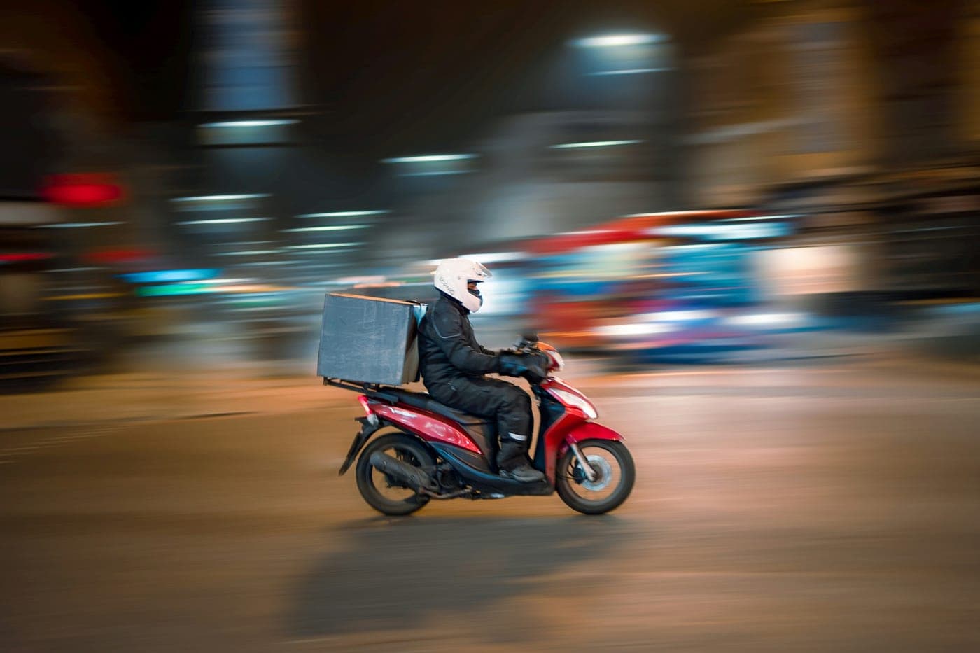 Entregador de moto com baú de entrega passando por uma rua iluminada à noite