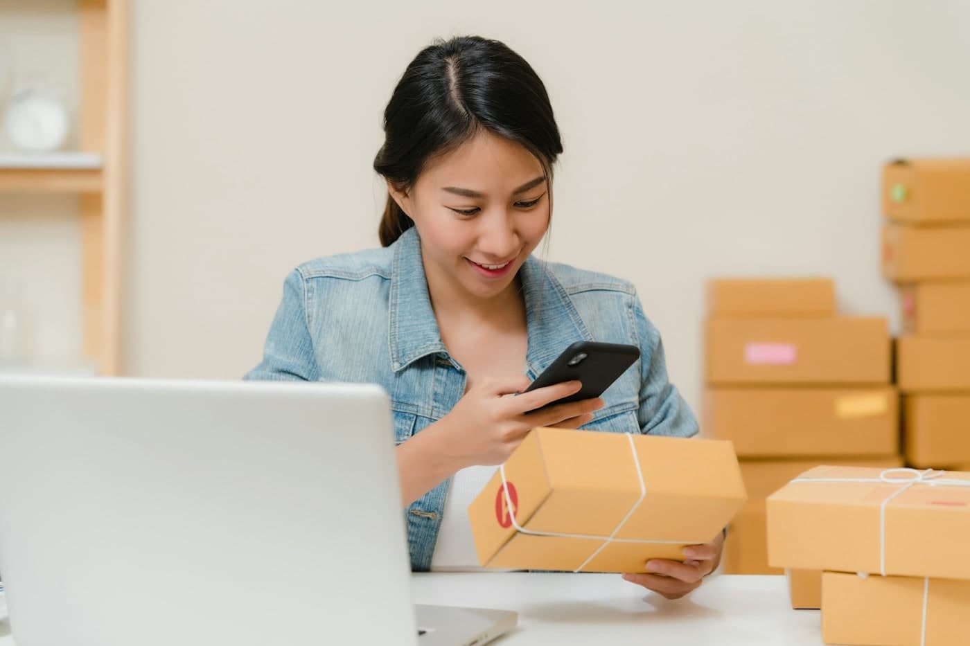 Lojista conferindo no celular um pedido embalado, com caixas de envio ao fundo